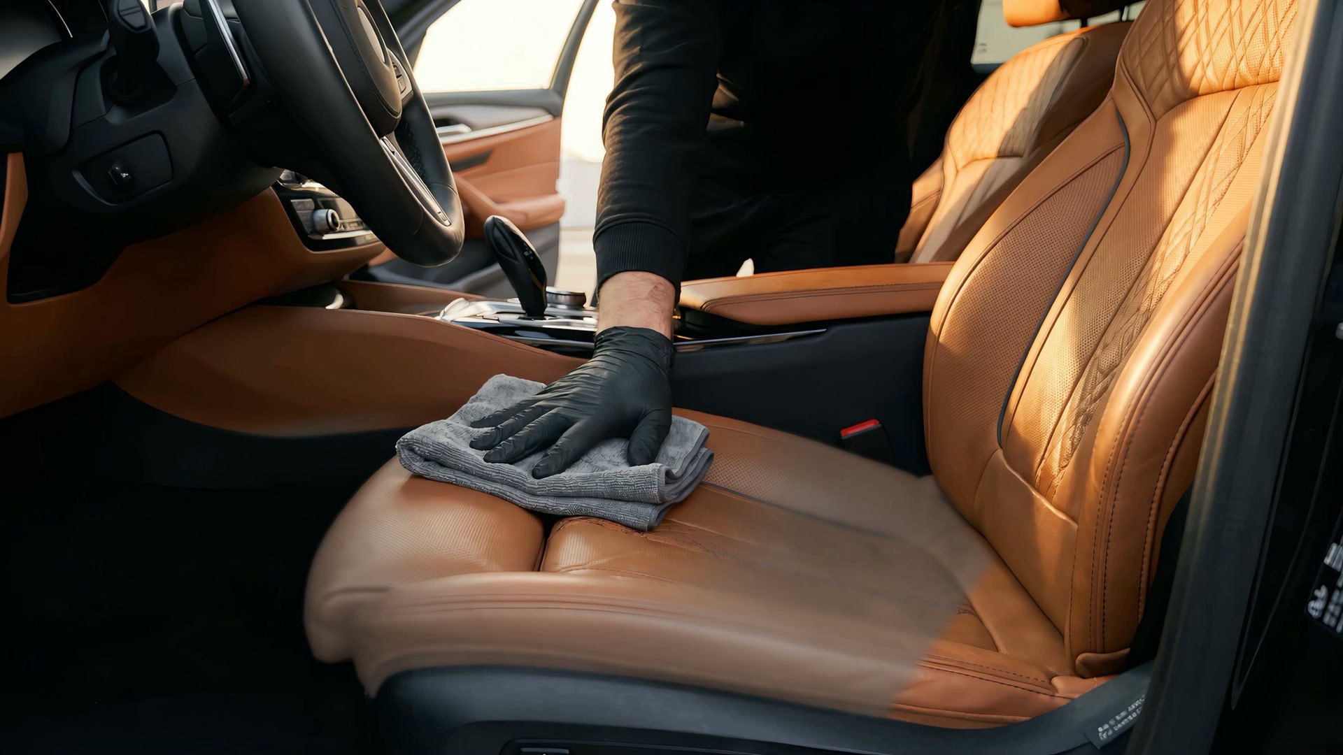 A black-gloved hand pressing a folded gray microfiber into a cognac quilted leather seat, the dark cabin and the steering wheel behind