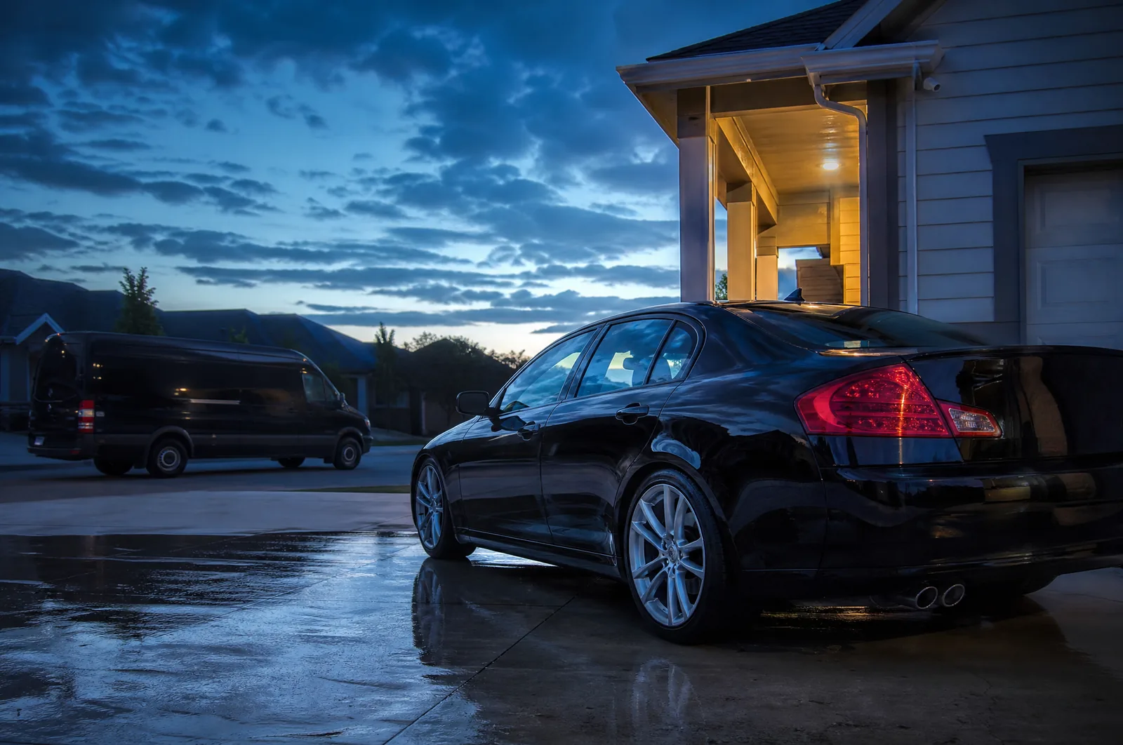 A freshly detailed black sedan on a wet driveway at blue hour, the detailing van parked in the background under a deep blue sky