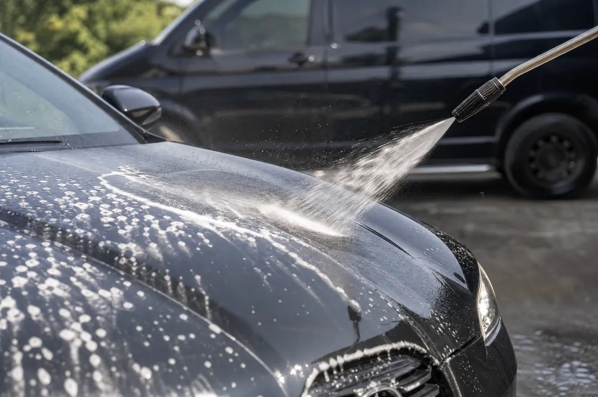 A pressure washer rinsing foam off a black hood on a driveway, the unmarked black van parked behind