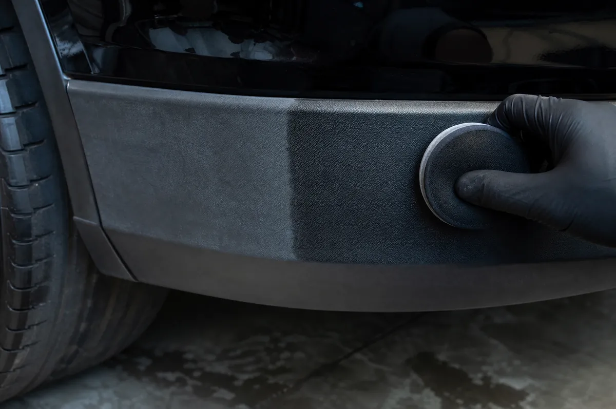 A gloved hand applying restorer to faded black plastic trim on a bumper